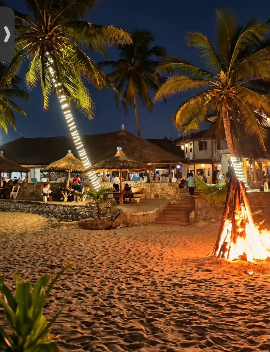 Evening bonfire on sand beside illuminated palms and resort seating